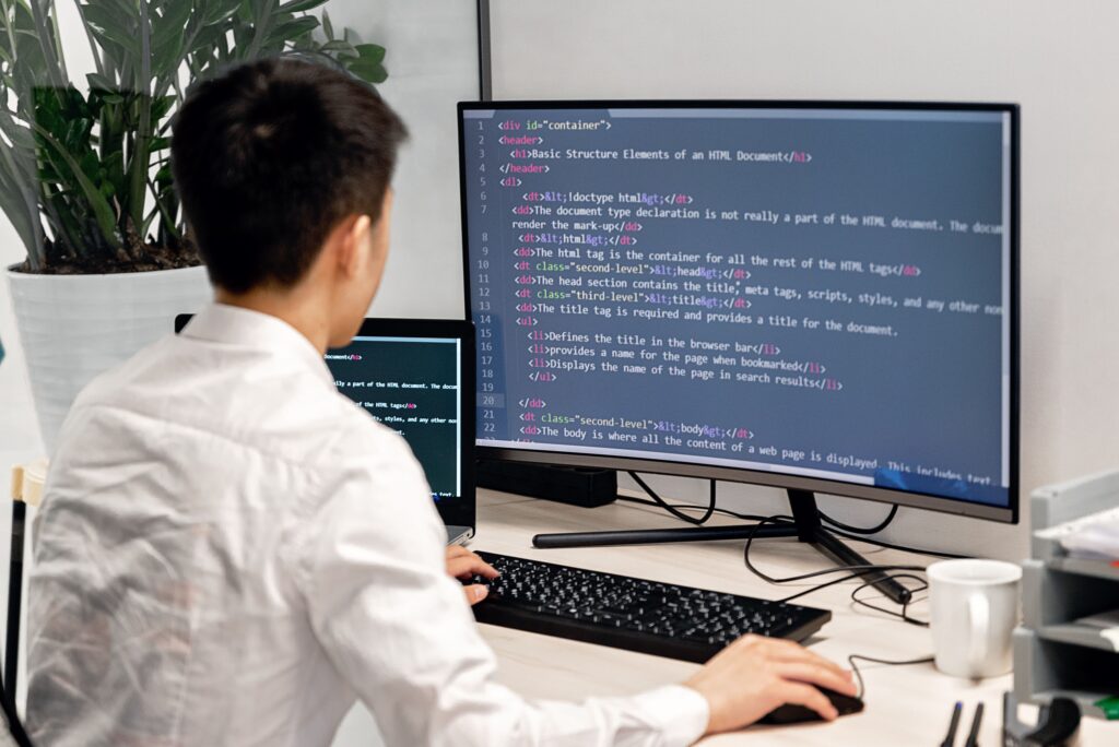 Adult male programmer coding on dual monitors in a modern indoor workspace.